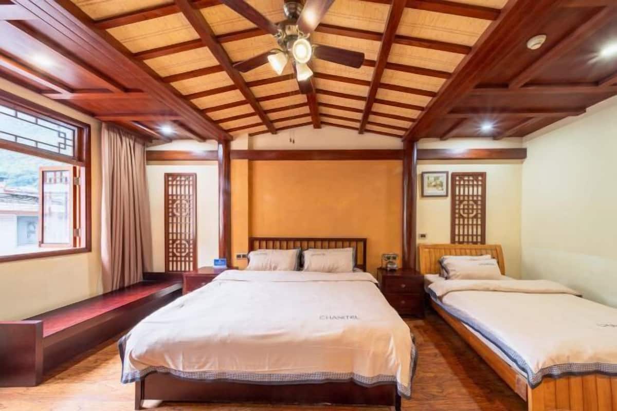 A hotel room with a large bed, a smaller bed, a ceiling fan, a window with curtains, and a wooden ceiling.