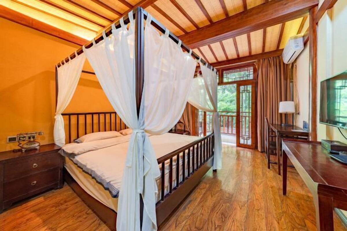 A four-poster bed with white drapes, a wooden floor, a desk, and a window with curtains.
