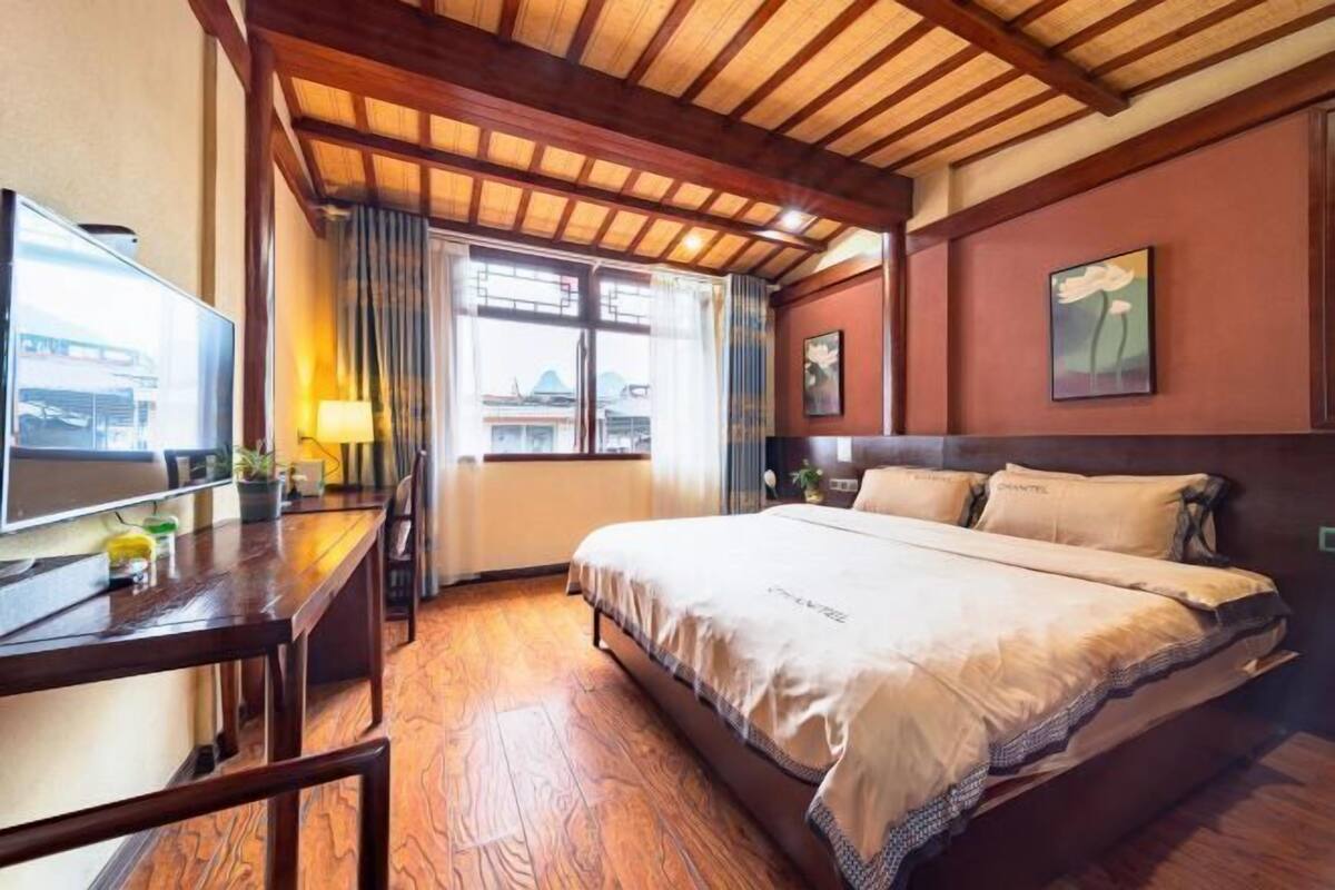 A hotel room with a wooden ceiling, a large bed with beige bedding, a desk with a lamp, and a view of the outside through a window.