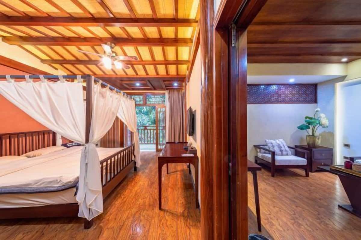 A room with a wooden ceiling, a bed with a canopy, a wooden table, a chair, and a small desk with a lamp.
