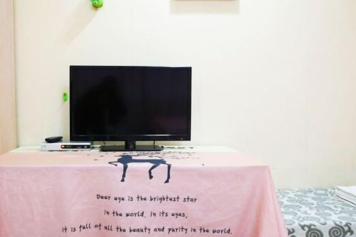 A room with a television on a table, a pink tablecloth with a quote, and a patterned bedspread.