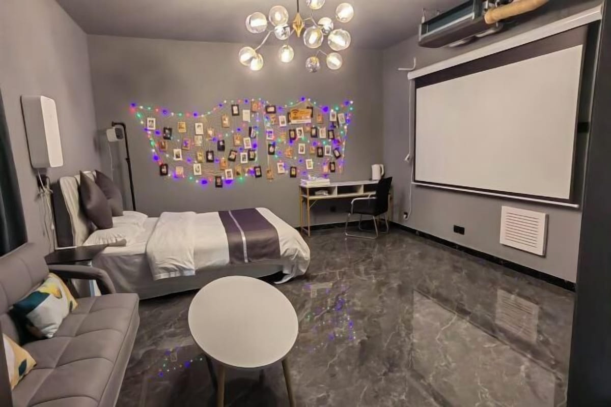 A modern hotel room with a bed, a sofa, a desk, and a projector screen.