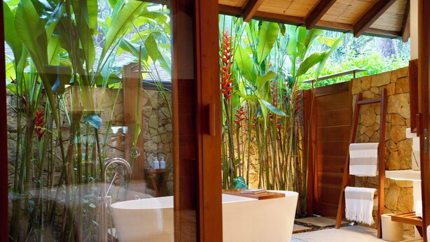Un baño moderno con una bañera independiente, paredes de piedra y exuberante follaje tropical.