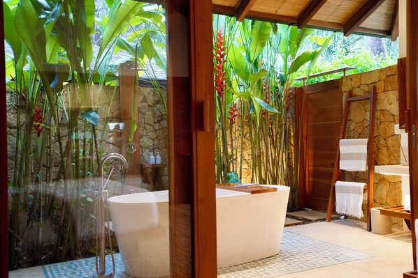 A modern bathroom with a freestanding bathtub, stone walls, and lush tropical plants.