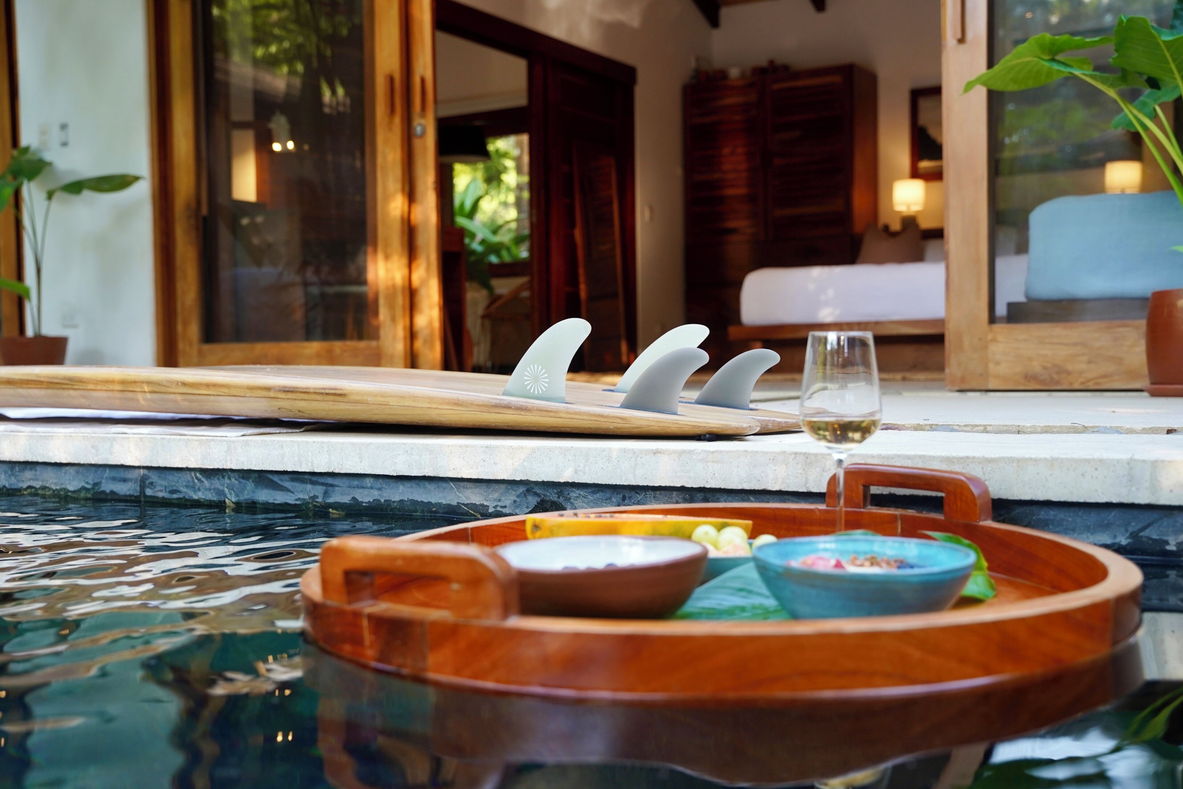 A wooden tray with bowls and a glass of white wine by a poolside.
