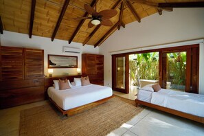 A bedroom with a large bed, a smaller bed, wooden walls, a ceiling fan, and a view of greenery outside.
