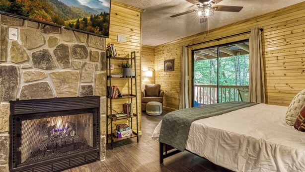 A cozy cabin room with a stone fireplace, a bed, a chair, and a balcony with a view of the forest.