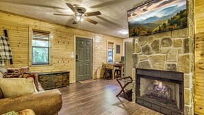 A log cabin interior with a fireplace, wooden walls, a dining area, and a ceiling fan.