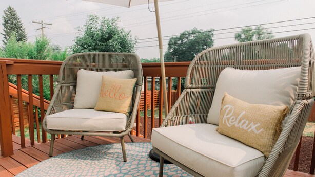 Twee rieten lounge fauteuils met kussens op een houten terras, een vloerkleed met patroon en een houten leuning.