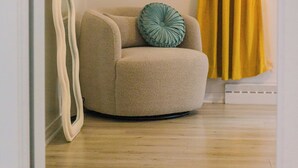 Een kamer met een beige fauteuil, een blauwgroen kussen, een geel gordijn en een wandplank met decoratieve objecten.