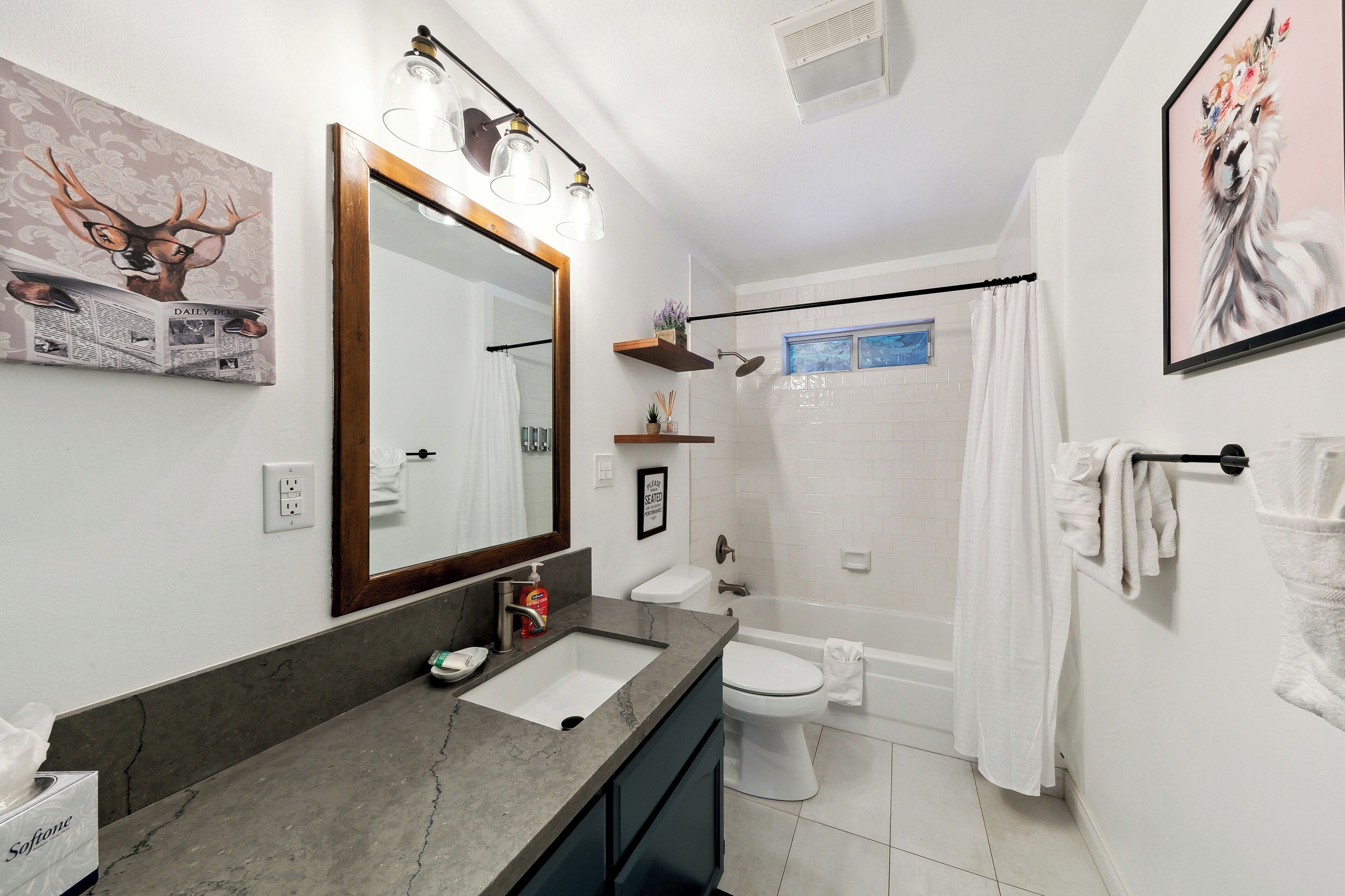 Ein Badezimmer mit Waschbecken, Toilette und Dusche. An den Wänden befinden sich Bilder in Rahmen, und über dem Waschbecken hängt ein Spiegel.