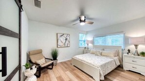 Un dormitorio con cama, una silla, una mesita de noche, ventilador de techo y una ventana con persianas.