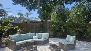 Un sofá seccional de mimbre con cojines verde claro y un sillón a juego, junto con dos mesas de centro de mimbre, sobre un patio de grava rodeado de exuberante follaje tropical y una cerca de madera.