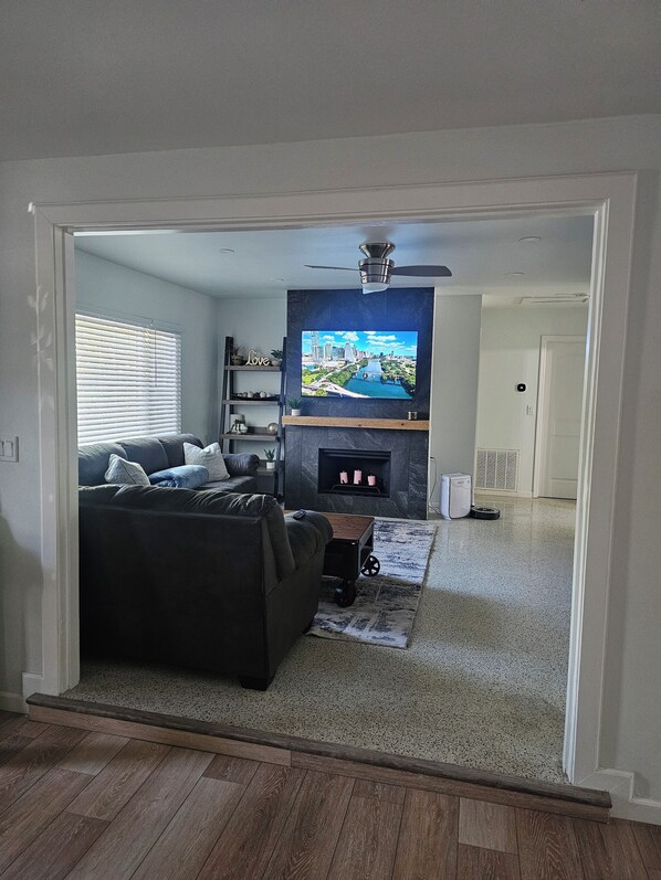 Ein Wohnzimmer mit einem dunkelgrauen Sofa, einem flachbildfernseher an der Wand, einem Deckenventilator und einem Kamin.