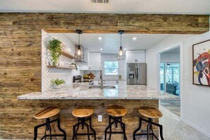 Una cocina con una pared decorativa de madera, una isla de granito con taburetes de bar, iluminación colgante y un refrigerador de acero inoxidable.