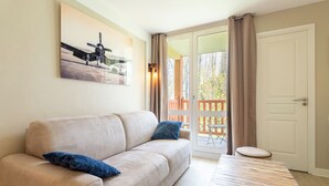 A living room with a beige sofa, two blue pillows, a wooden coffee table, and a painting of an airplane on the wall. - Appt 2 pièces cabine 6 pers avec piscine, tennis, parking gratuit 157 (Le Touquet-Paris-Plage)