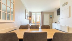 A dining area with a wooden table and six chairs, a kitchen area with cabinets and a TV, and a view of a balcony with curtains. - Appt 2 pièces cabine 6 pers avec piscine, tennis, parking gratuit 157 (Le Touquet-Paris-Plage)
