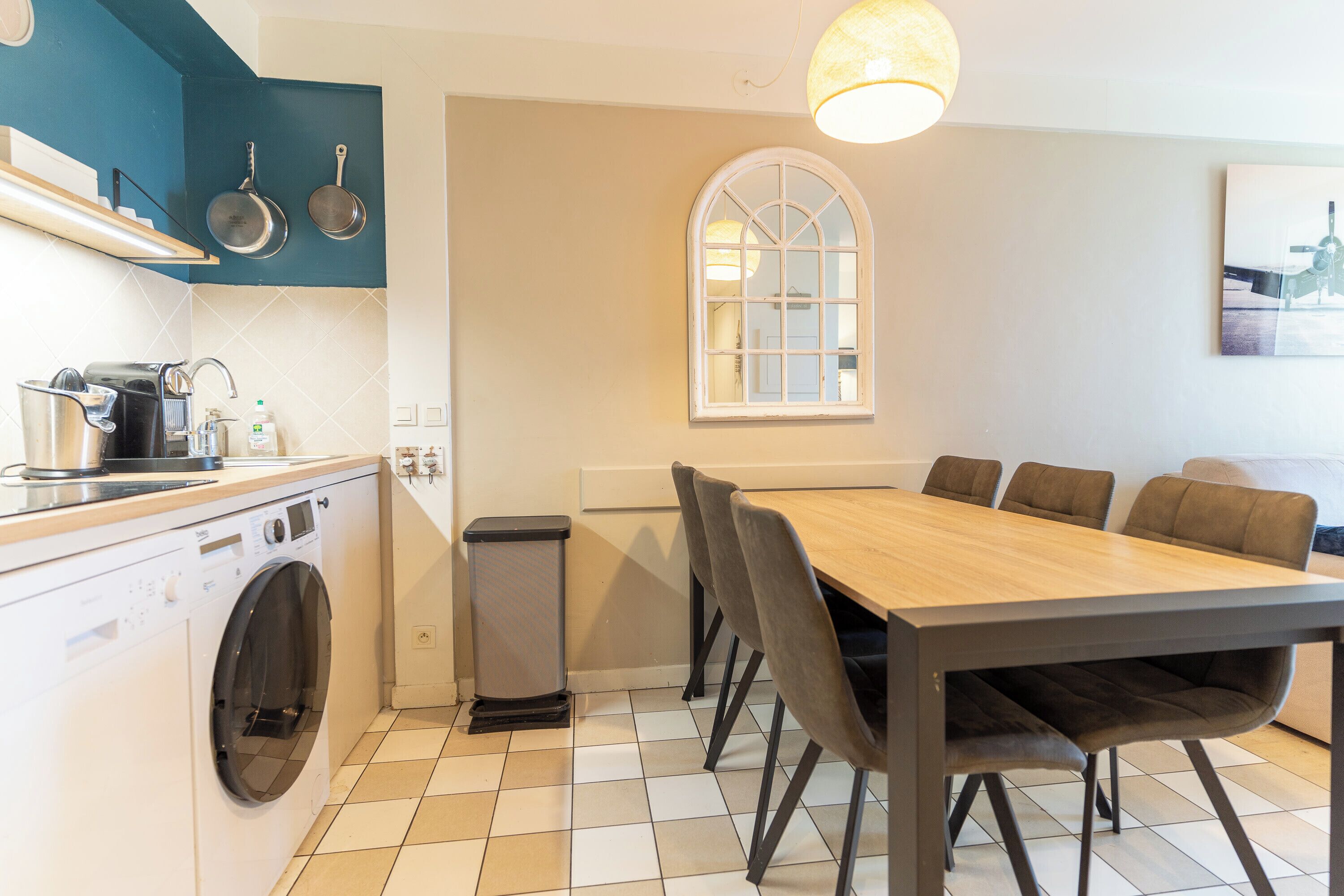 A compact kitchen with a washing machine, a dining table with chairs, and a window with a view.
