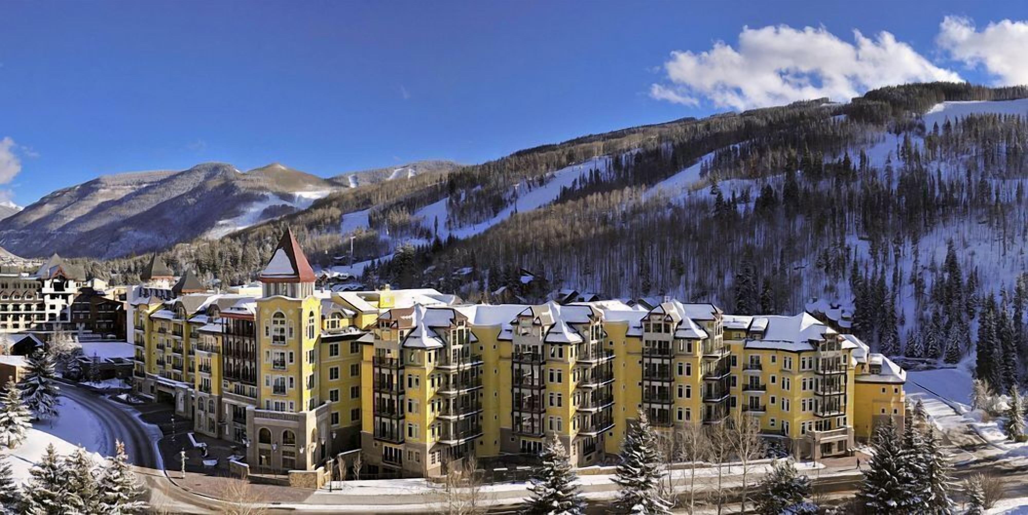 A multi-story hotel with a central tower, surrounded by snow-covered mountains and a clear sky.