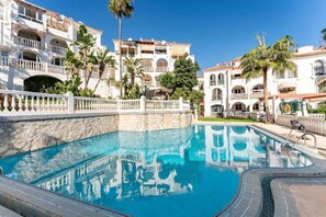 A swimming pool with a stone wall, surrounded by multi-story buildings with balconies and palm trees.