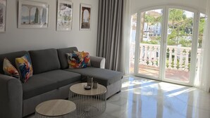 Una sala de estar moderna con un sofá gris, dos mesitas redondas y un amplio ventanal corredera que da a un balcón.