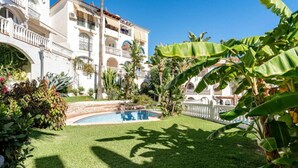 Una piscina rodeada de un césped cuidado, con un edificio al fondo que tiene balcones y un muro de piedra.