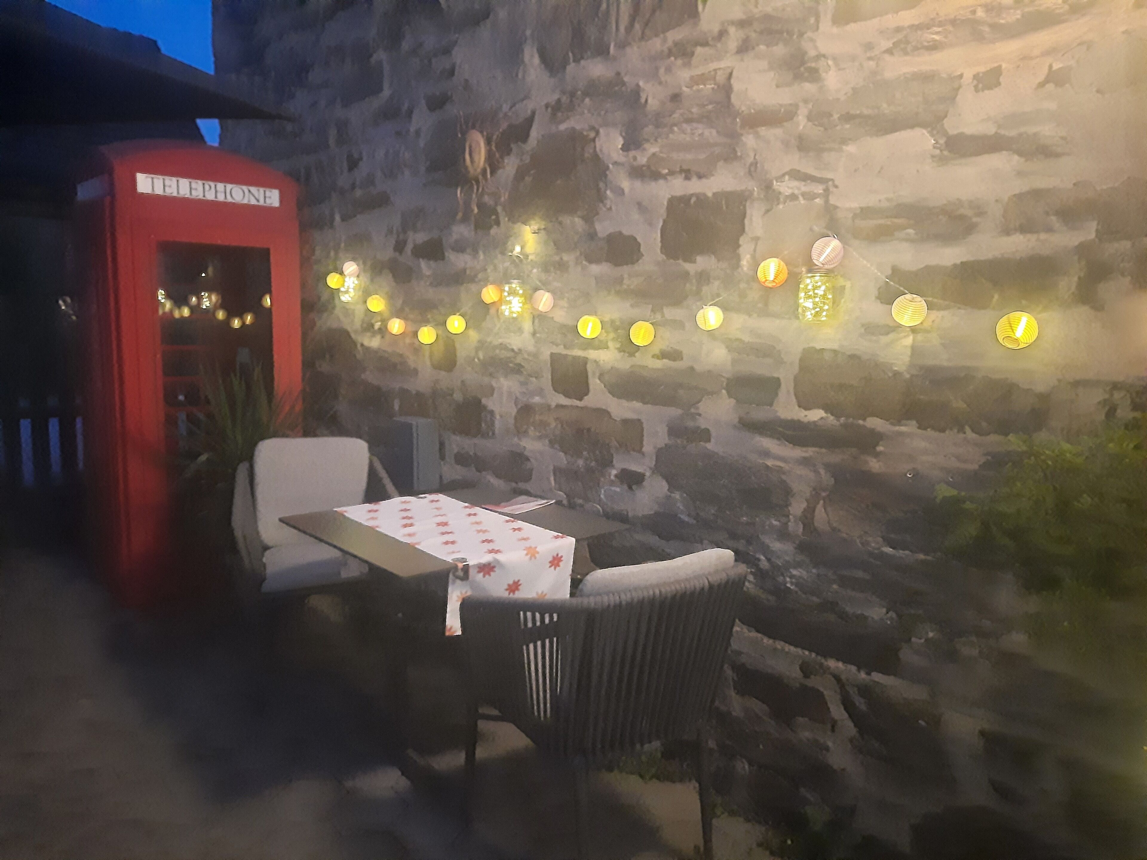 Un mur de pierre avec une cabine téléphonique, des sièges extérieurs avec une table et des chaises, et des guirlandes lumineuses.