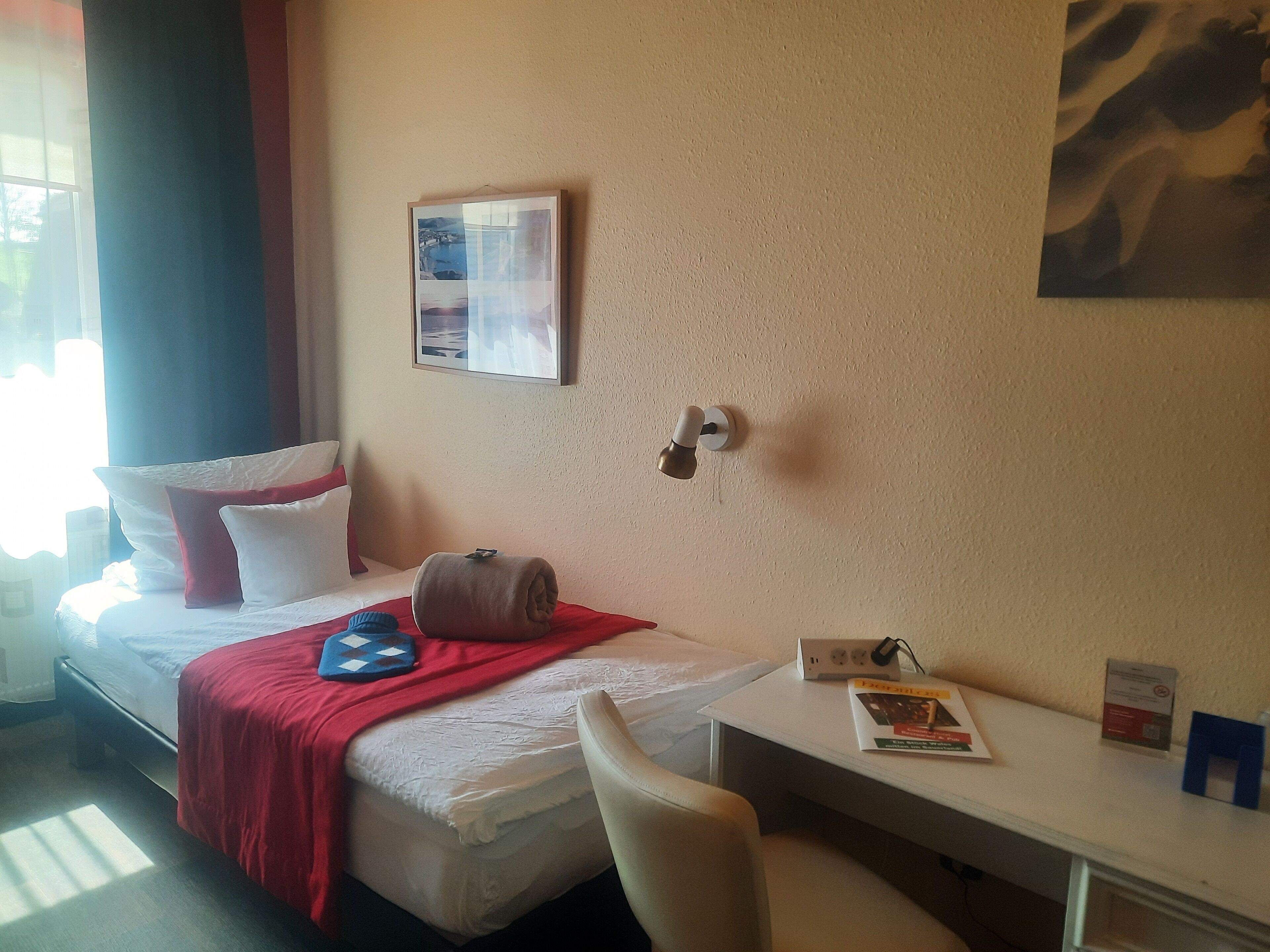 A hotel room with a bed, a desk, a chair, a wall-mounted picture, and a door lock.