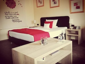 A hotel room with a bed, bedside tables, a wooden nightstand, a wall with a quote, and a small table with a vase. - Henblas-Hotel (Neuenrade)