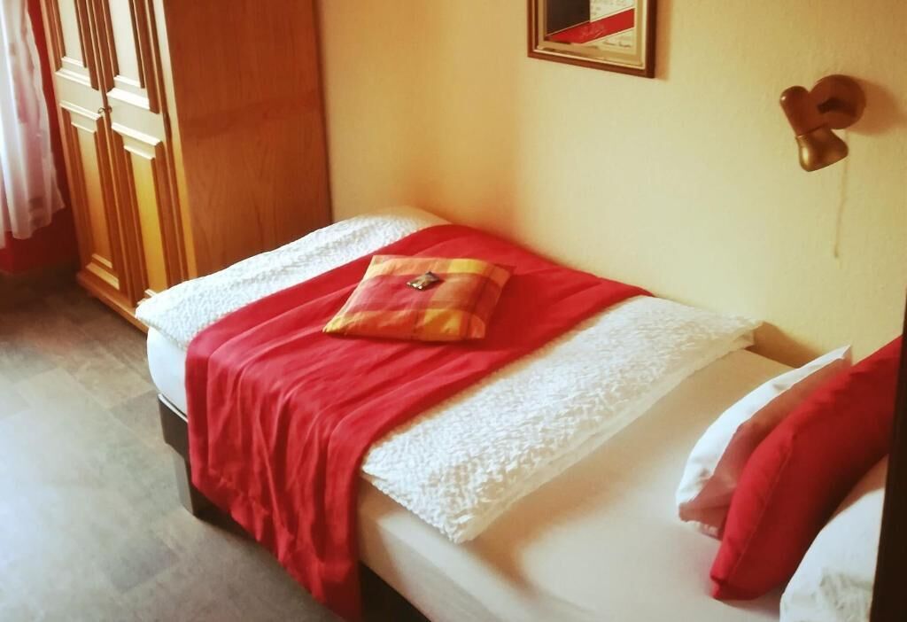 Una cama individual con una colcha roja y un almohadón, un armario de madera y un objeto con forma de corazón en la pared.