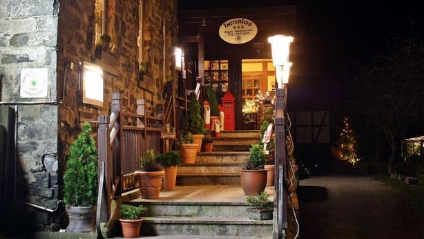 A stone building with a sign reading "henblas" and a red door. There are steps leading to the entrance, potted plants, and a Christmas tree in the background. - Henblas-Hotel (Neuenrade)