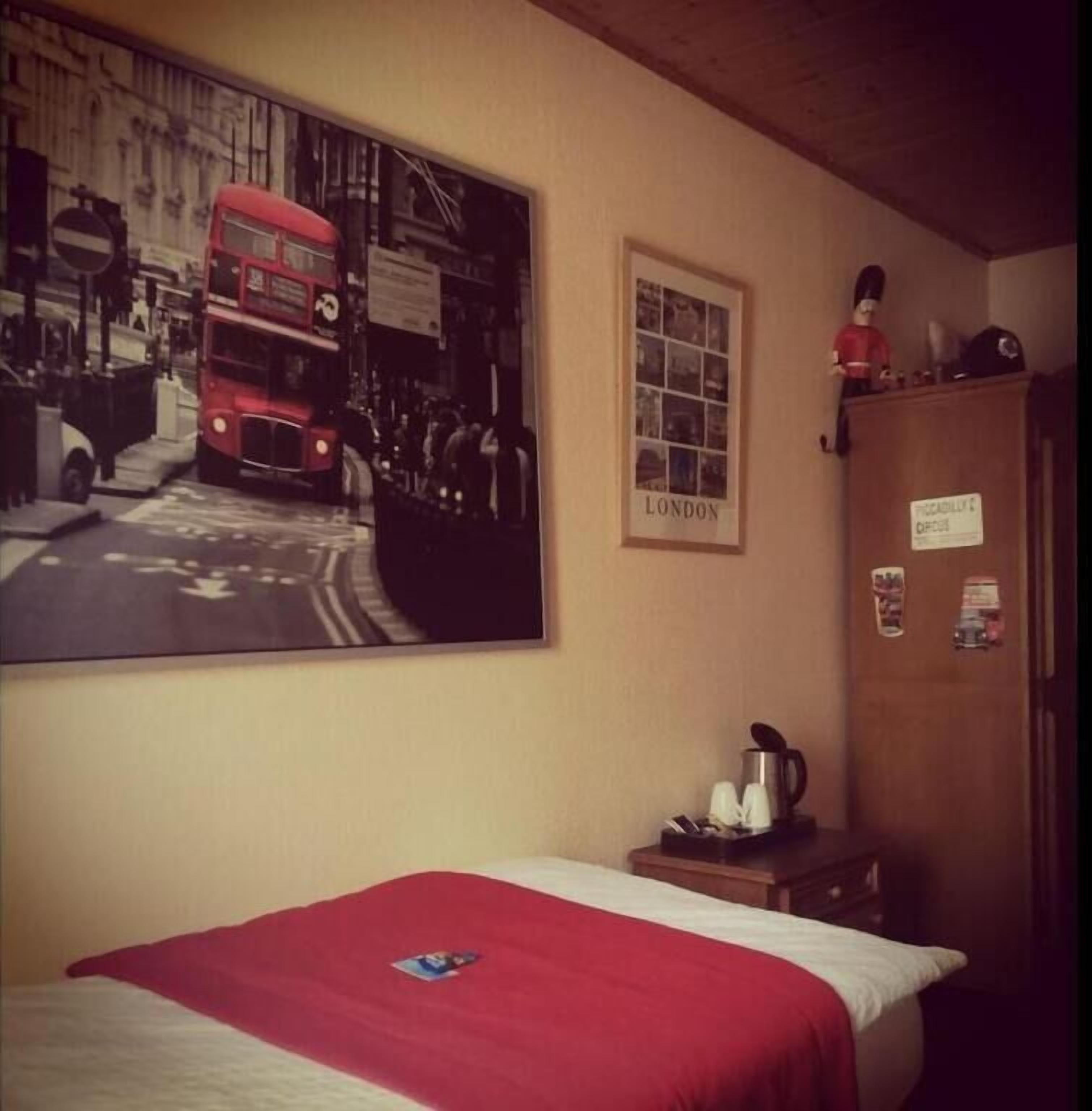 A hotel room with a bed, a large poster of a red double-decker bus, a framed picture of London, and a wooden wardrobe.