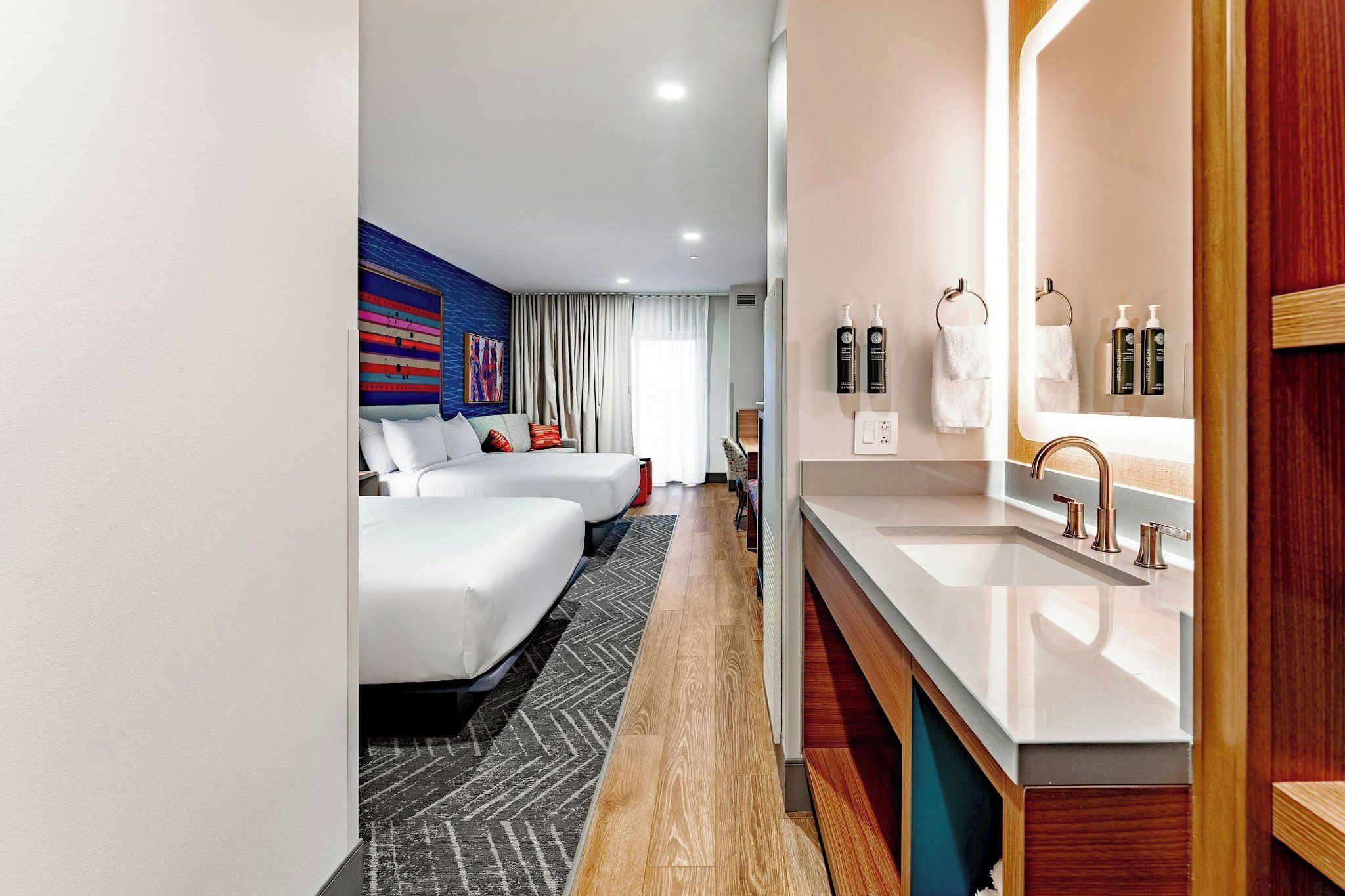 A hotel room with two beds, a large mirror, a vanity with a sink, and a view of the hallway.