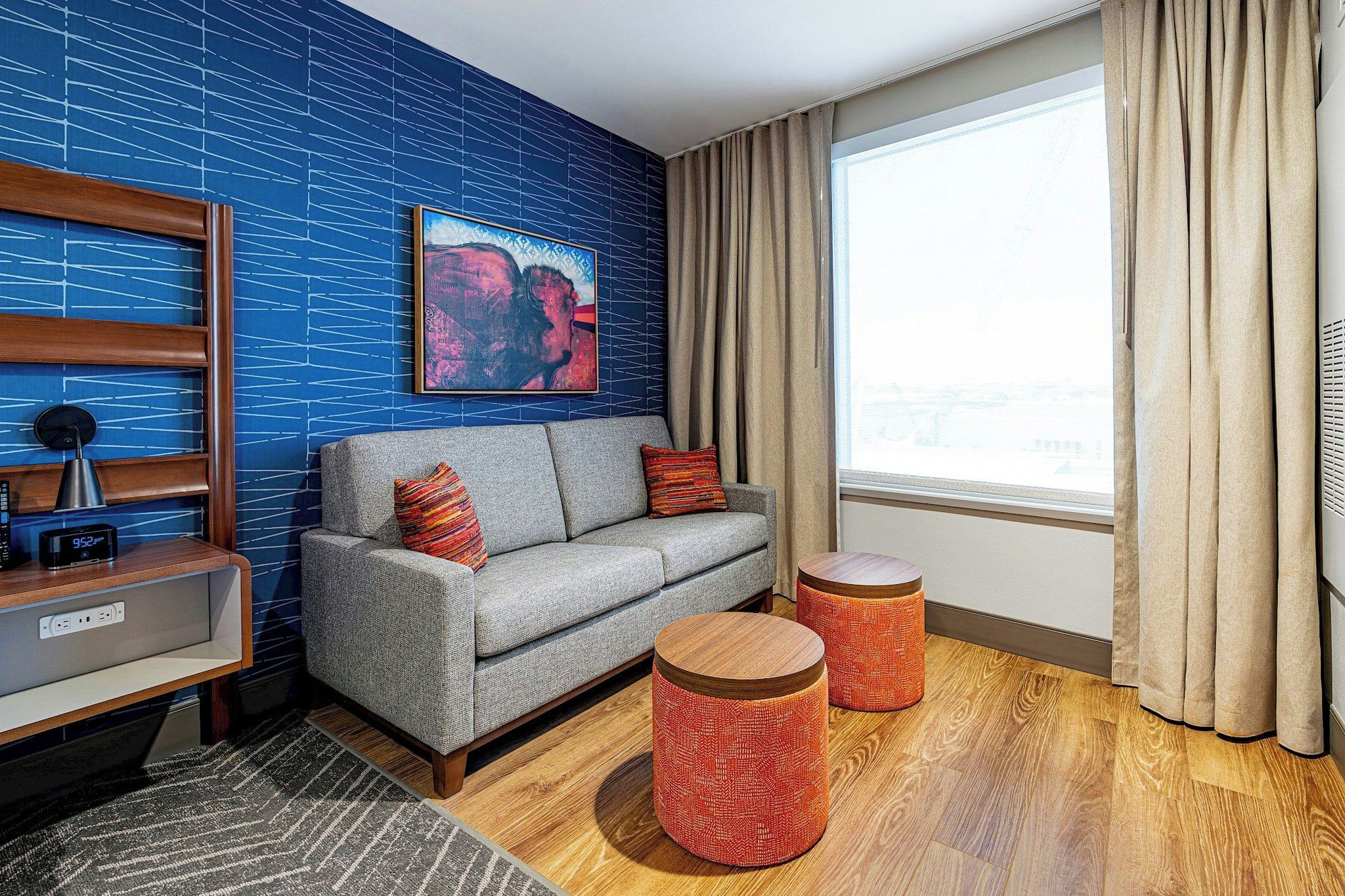 A modern living room with a grey sofa, two red patterned ottomans, a blue patterned wall, and a window with beige curtains.