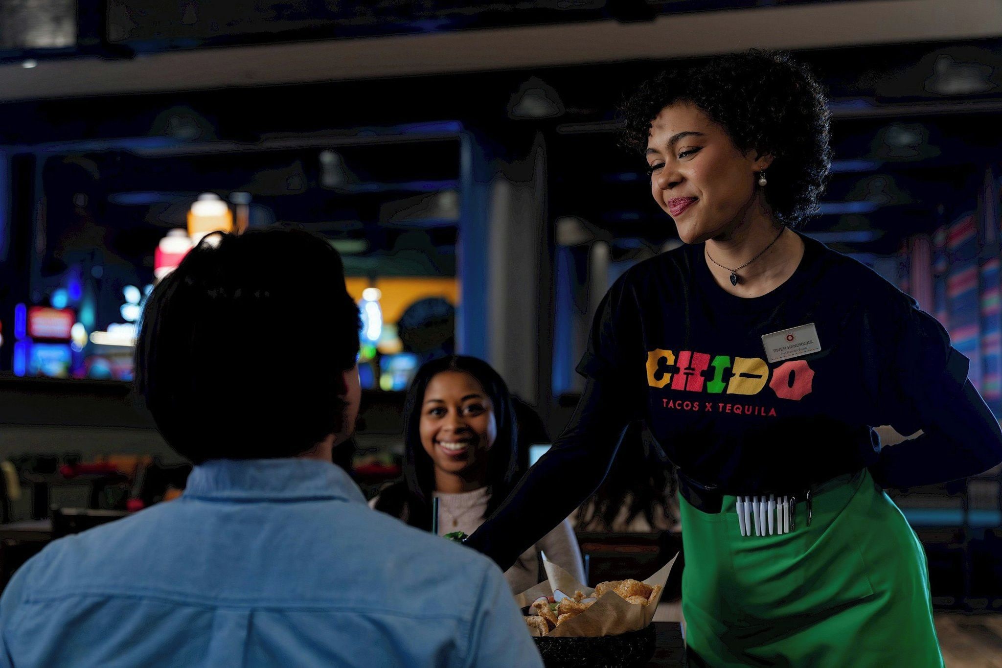 A person wearing a name tag and a shirt with a colorful design, standing in a dimly lit indoor setting.