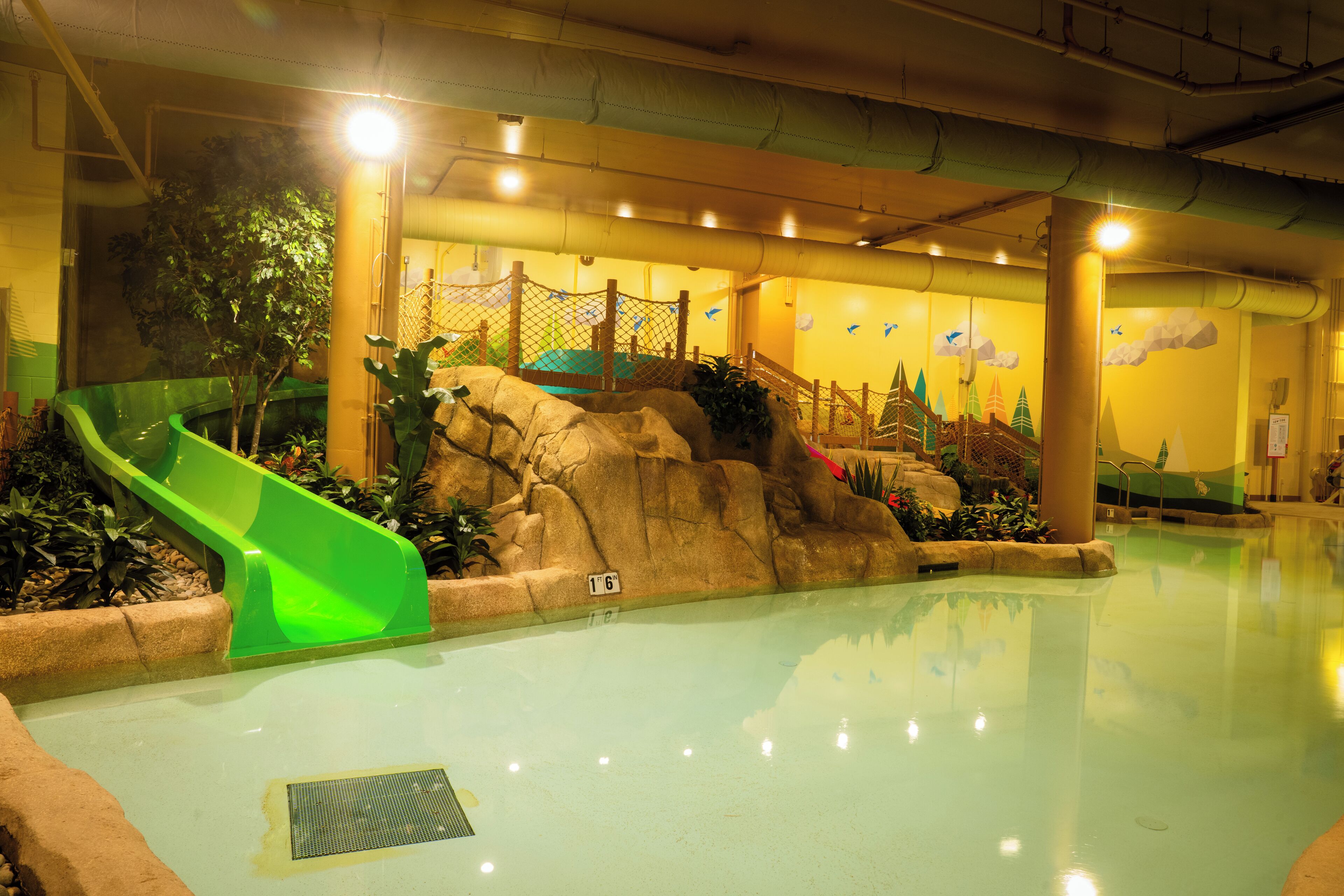 Una piscina interior con un tobogán verde, formaciones rocosas y un mural de un castillo en la pared.