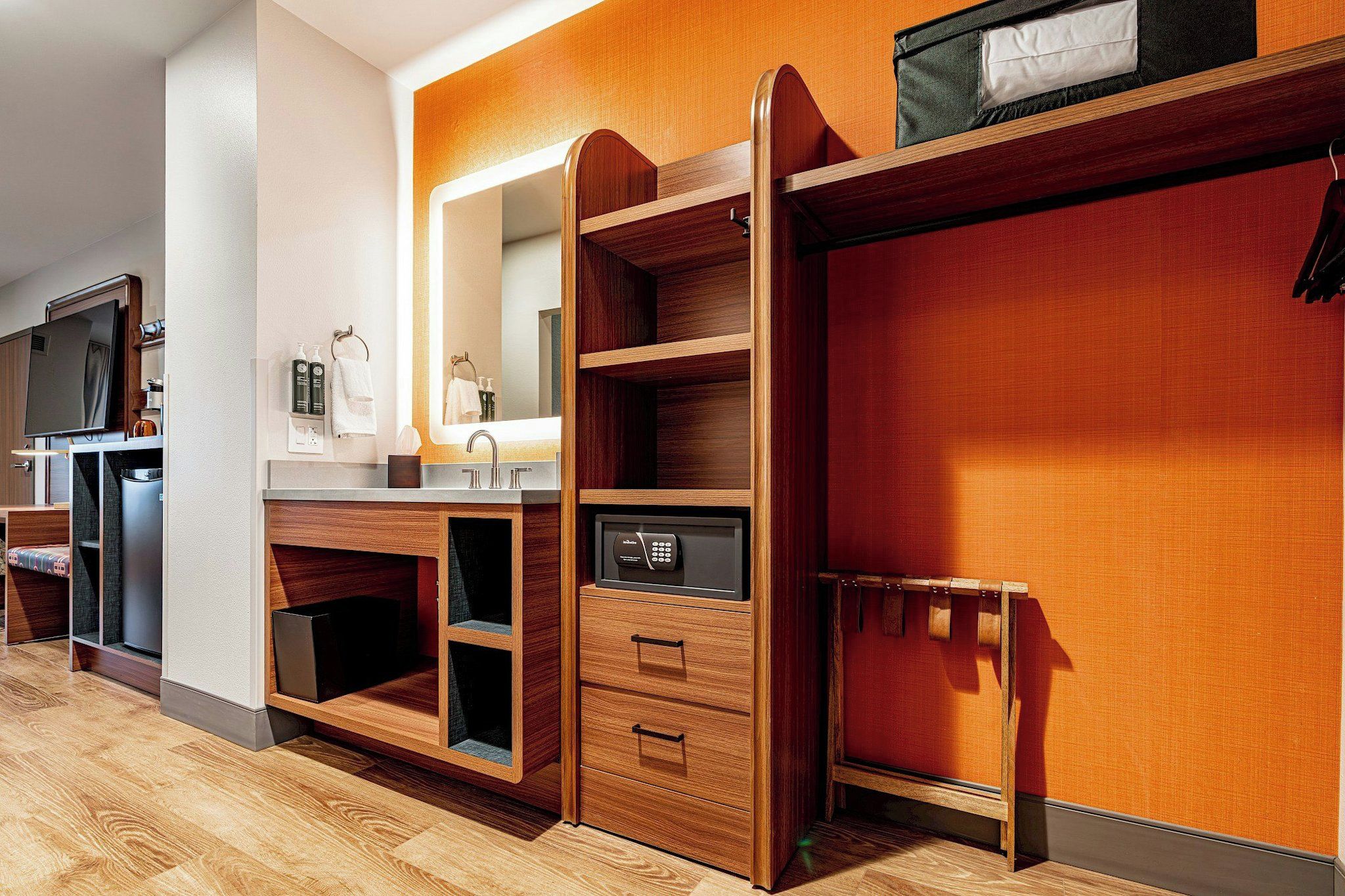 A compact hotel room with a wooden vanity, a built-in TV, and a small kitchenette.