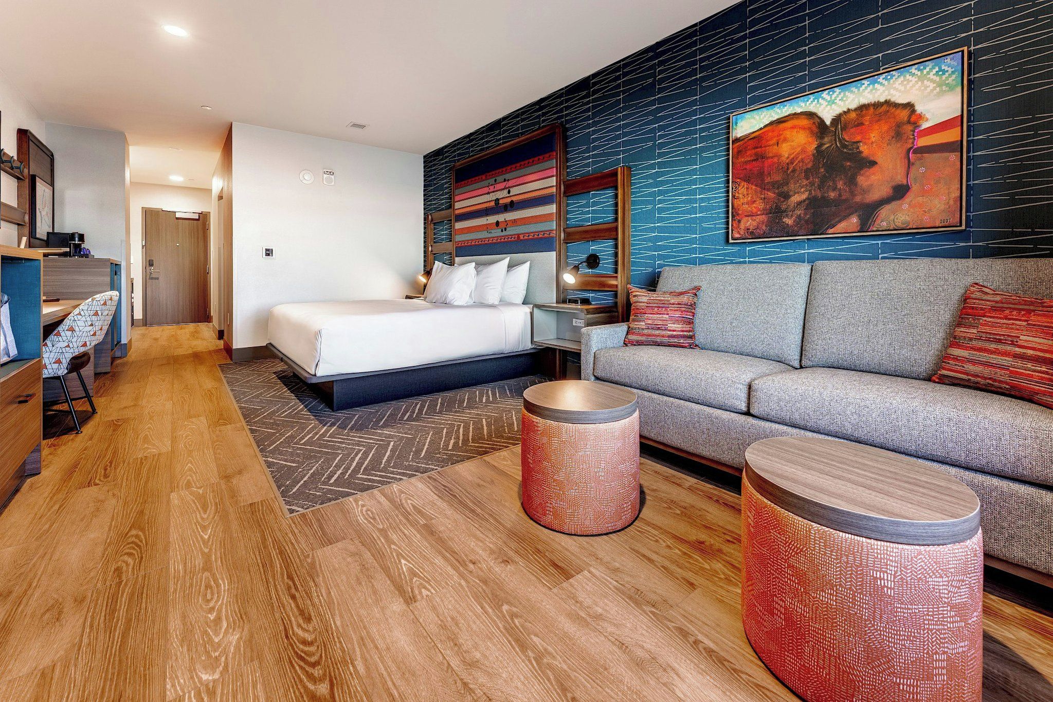 A modern hotel room with a bed, a sofa, two round side tables, a desk with a chair, and a large abstract painting on the wall.