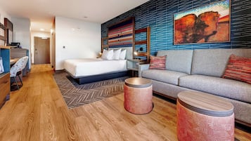 A modern hotel room with a bed, a sofa, two round side tables, a desk with a chair, and a large abstract painting on the wall.