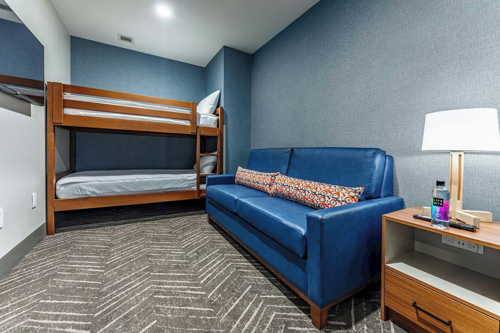 Una habitación con una cama litera, un sofá azul, un cojín estampado, una lámpara, una botella y un control remoto sobre una mesita de noche de madera.