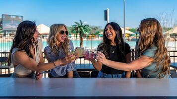 Four people toasting with drinks at an outdoor table with a pool in the background.