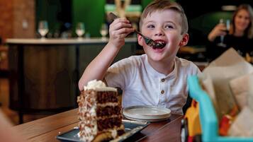 Ein Kind isst mit einem Löffel Kuchen an einem Holztisch, auf dem ein Teller und eine Kuchengabel liegen.