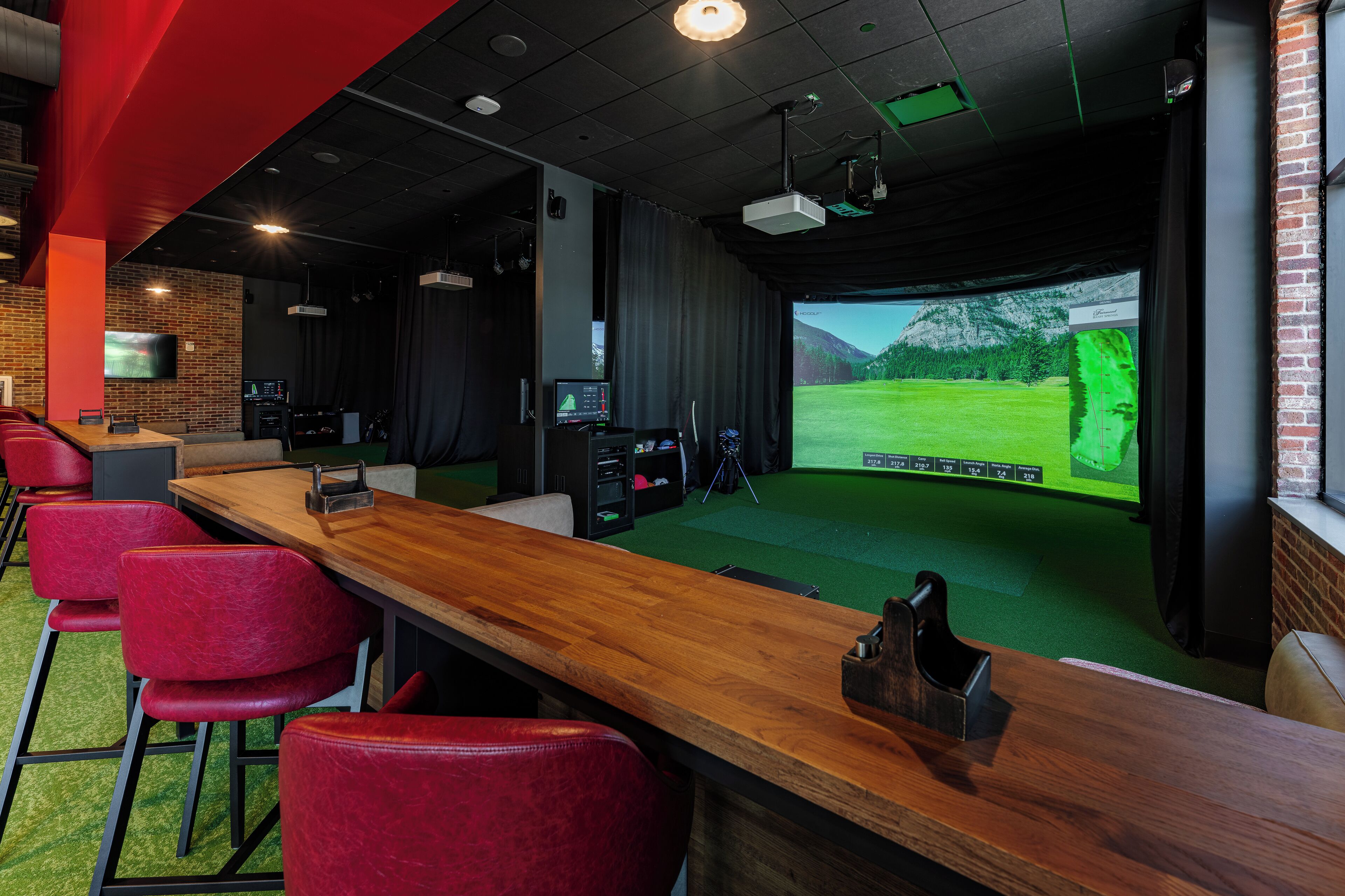Et værelse med et langt træbord, røde stole, et projektorskærm og en golfsimulator.