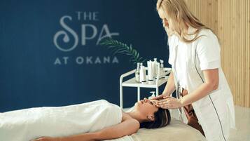 A spa treatment room with a client lying on a treatment table, a practitioner attending to the client, and a wall with the spa's name.