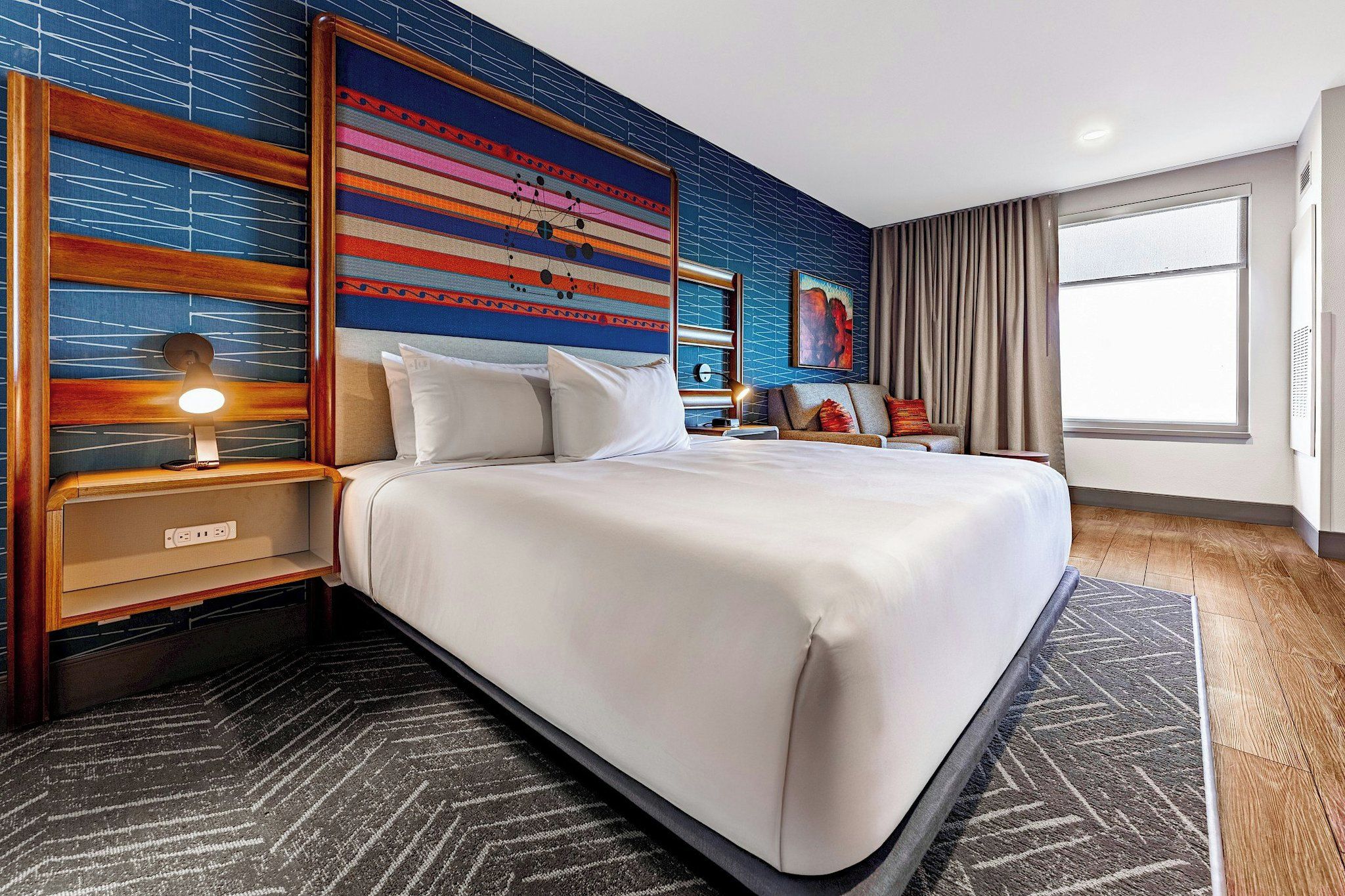 Une chambre d’hôtel avec un grand lit, une table de chevet avec une lampe, un mur orné d’un motif abstrait et coloré, et une fenêtre avec des rideaux.