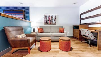A modern living room with a brown leather armchair, a grey sofa, two wooden side tables, and a wooden desk with a chair.