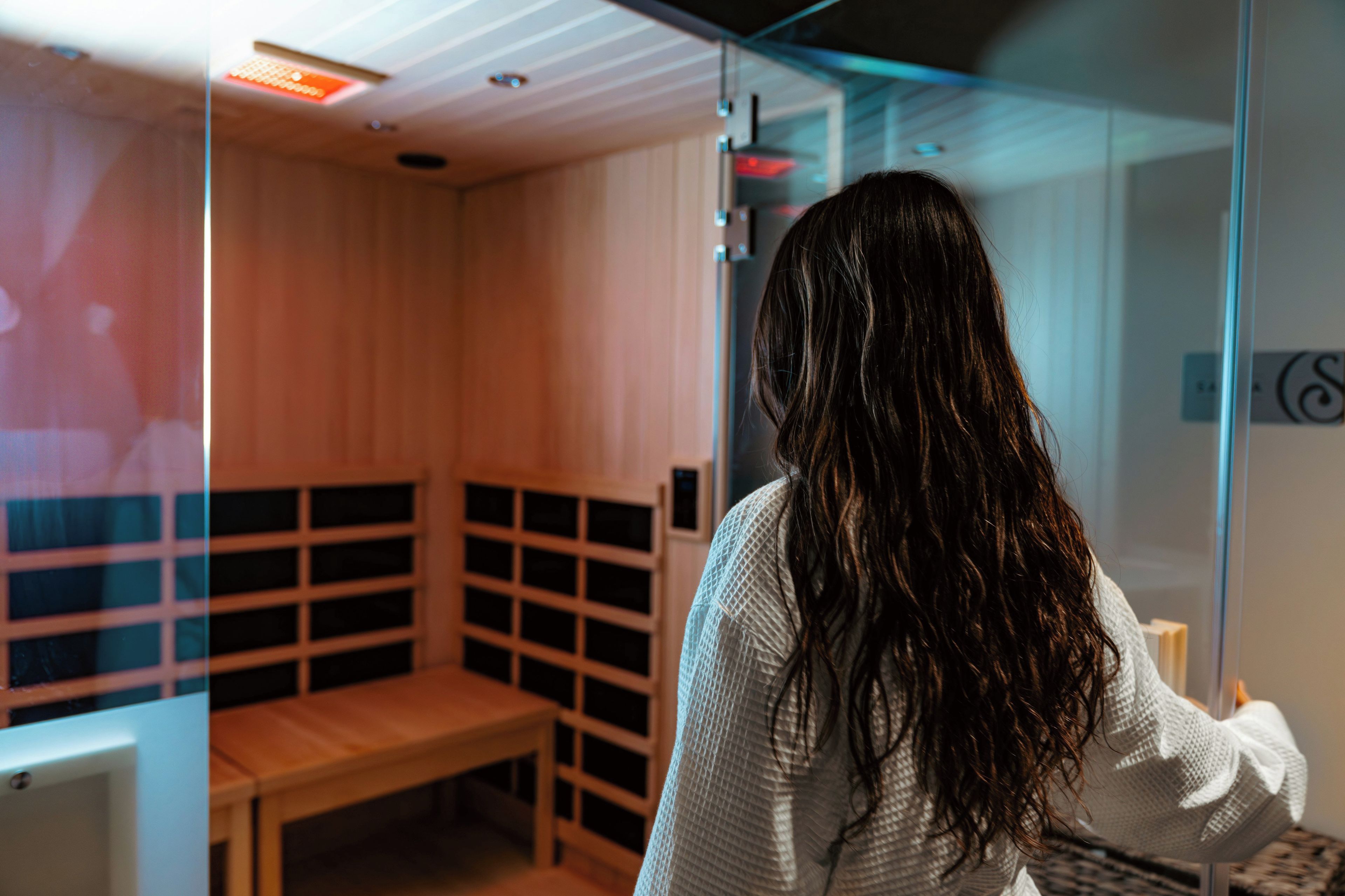 A person standing in a sauna room with a bench and a glass partition.