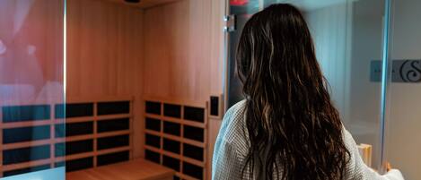 A person standing in a sauna room with a bench and a glass partition.