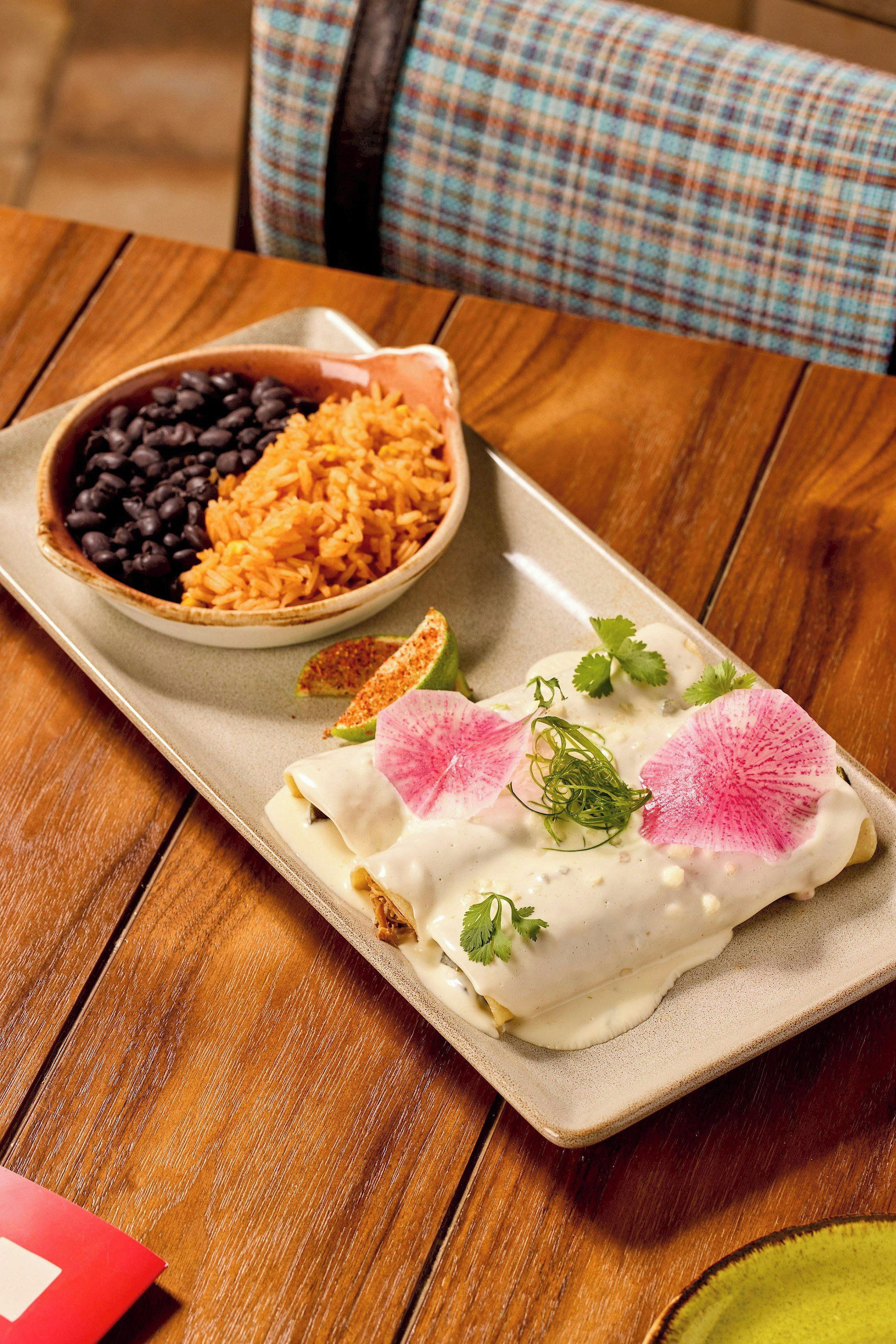A plate of enchiladas with a creamy sauce, garnished with radish slices and cilantro, served with a side of black beans and rice.