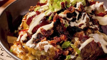 A bowl of nachos topped with cheese, bacon, and green onions, with a drink in the background.